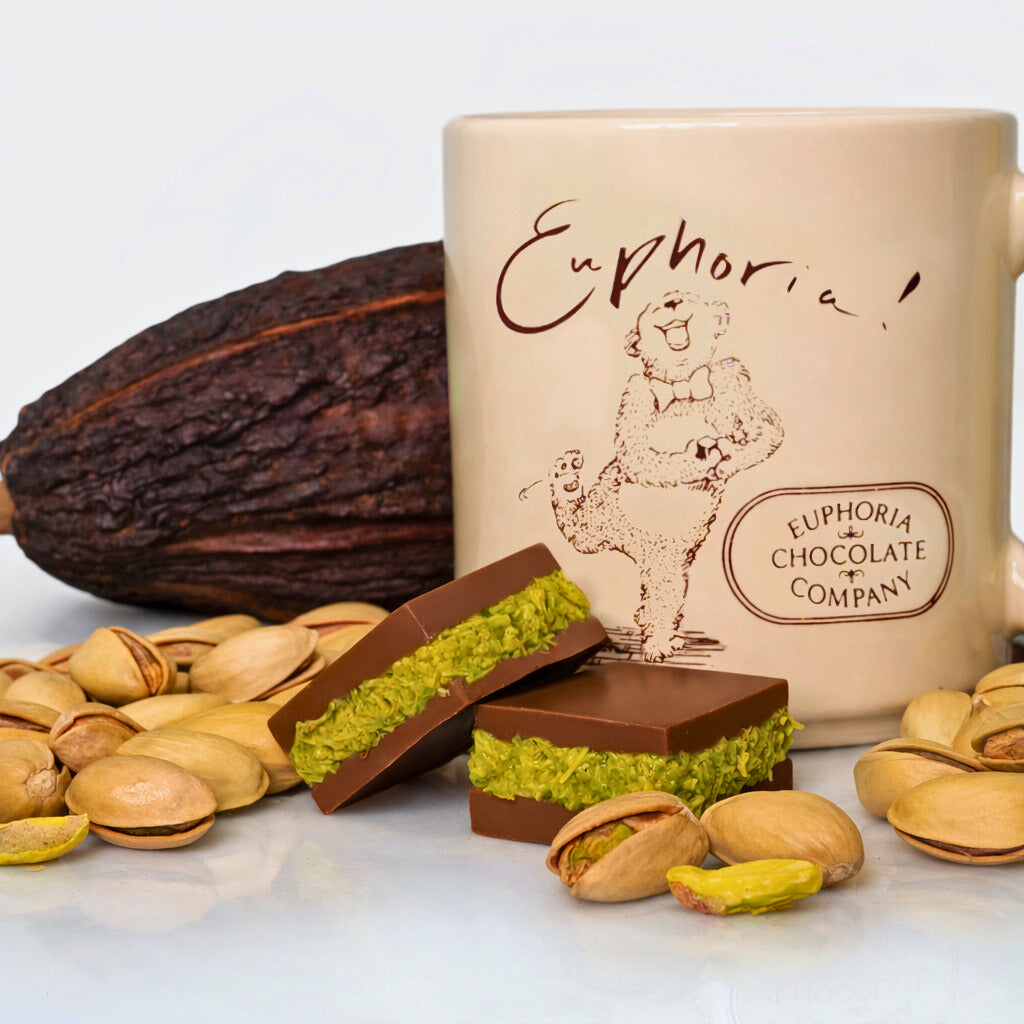 Mug with 'Euphoria Chocolate Company' branding, surrounded by chocolate bars, pistachios, and a cacao pod.