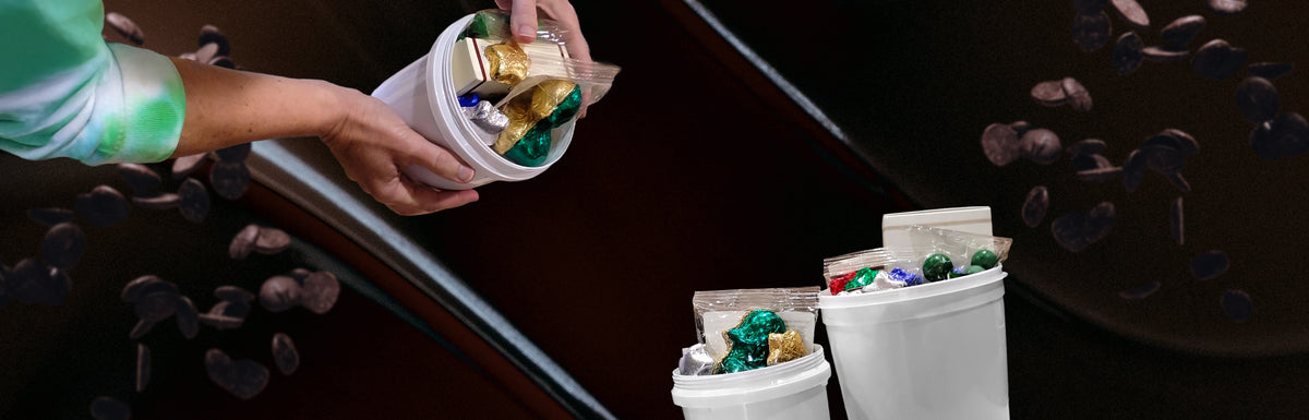 A person's hands holding a white plastic tub filled with wrapped chocolates, with more white plastic tubs containing assorted chocolates in the foreground and scattered chocolate pieces on a dark background.