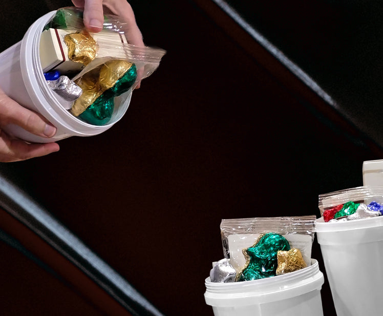 A close-up of a hand holding a white plastic tub filled with assorted foil-wrapped chocolates, with additional similar tubs visible in the background.
