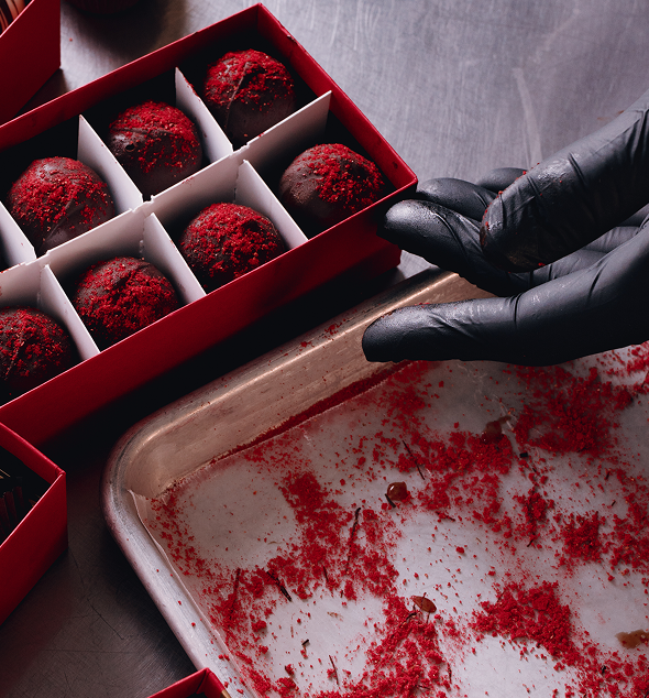 Chocolate truffles coated in red powder, shown in a red gift box next to a gloved hand reaching toward them on a dark surface.