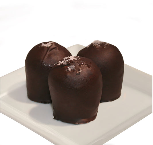 Three dark chocolate truffles with salt flakes on a white plate against a white background.