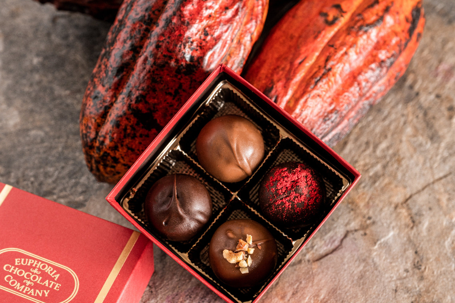 Box of chocolate truffles filled with various toppings with cacao pods on a stone surface