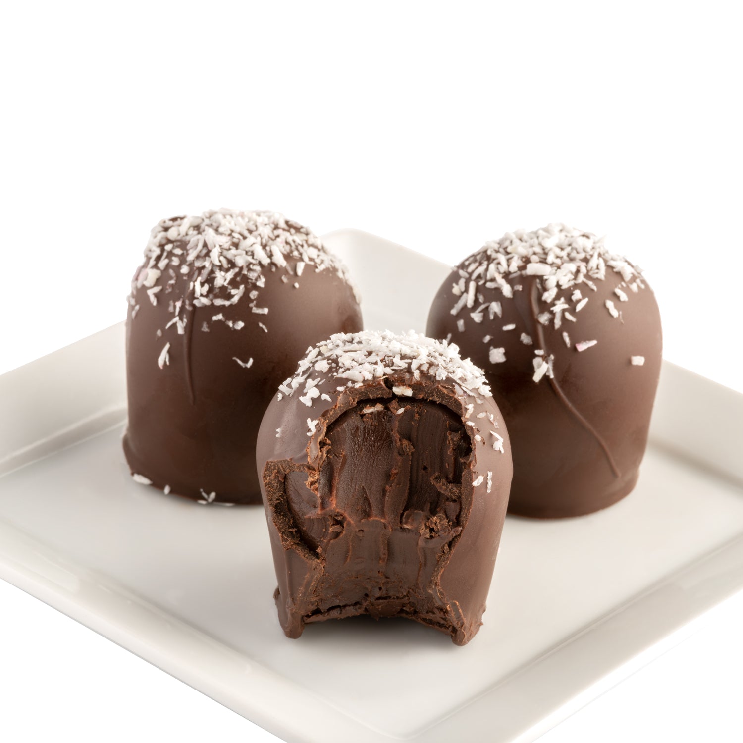 Three dark chocolate truffles with shredded coconut on top sitting on a white plate. One truffle has a bite taken out of it to show the dark chocolate core.