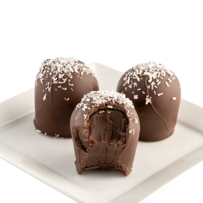 Three dark chocolate truffles with shredded coconut on top sitting on a white plate. One truffle has a bite taken out of it to show the dark chocolate core.
