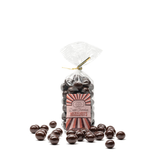 A clear plastic bag filled with dark chocolate covered hazelnuts, with a label showing the Euphoria Chocolate Company brand and the product name. A few chocolate-covered hazelnuts are scattered in front of the bag.