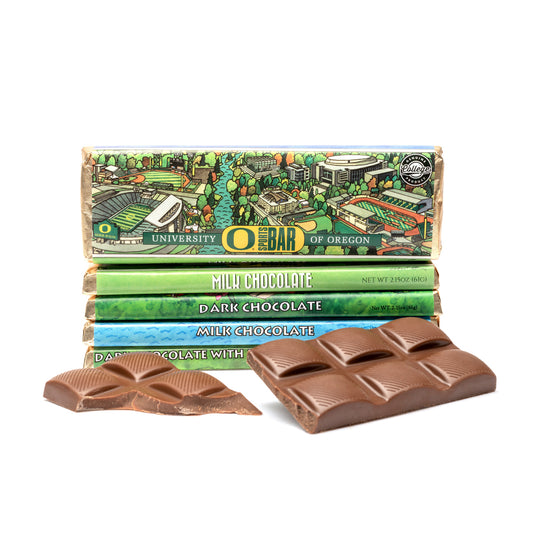 A milk chocolate bar with a wrapper featuring a hand-painted representation of the University of Oregon campus, along with pieces of broken chocolate showing the texture and color of the bar in front of a white background.