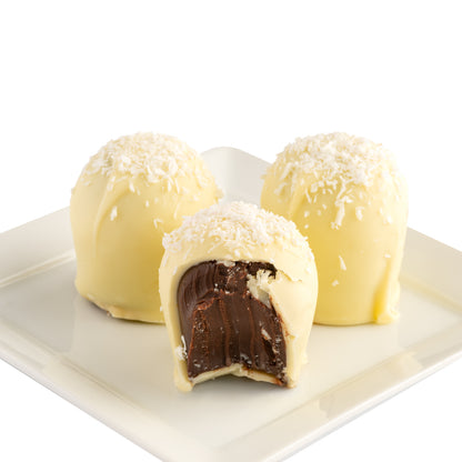 Three white chocolate truffles on a plate with shredded coconut on top; one has a bite taken out of it to expose the dark chocolate interior.