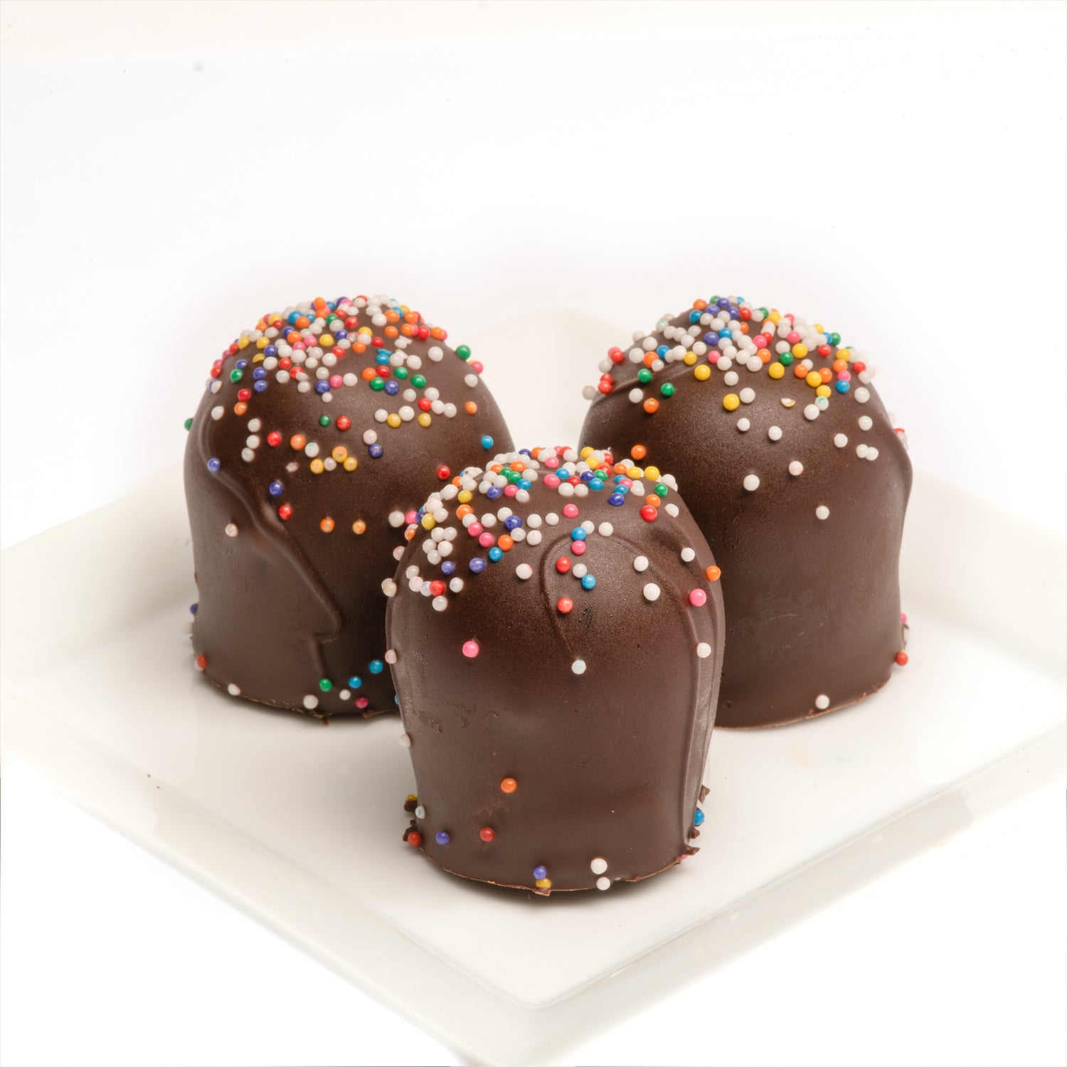 A plate of three hand-dipped chocolate birthday cake truffles with rainbow nonpareils on top in front of a white background.